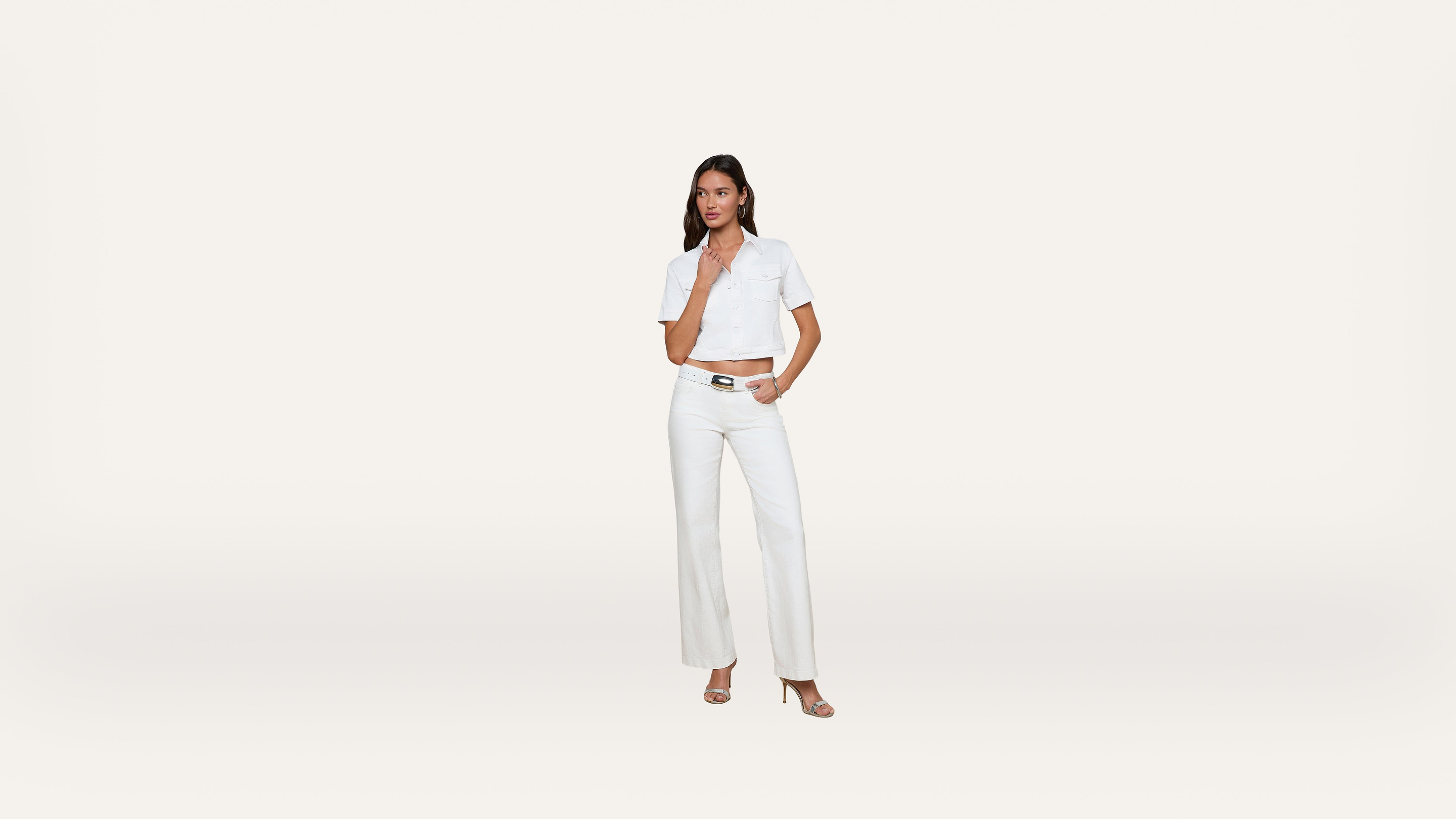 A woman stands against a plain white background, wearing a short-sleeved white button-up shirt, white high-waisted pants, and heeled sandals. She has one hand on her hip and the other touching her chin.