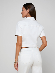 A woman stands facing away, wearing the Kirstie Jean Shirt Jacket with short sleeves and a pair of white jeans. She has long, straight hair and accessorizes with hoop earrings and bracelets against a plain, light-colored background.