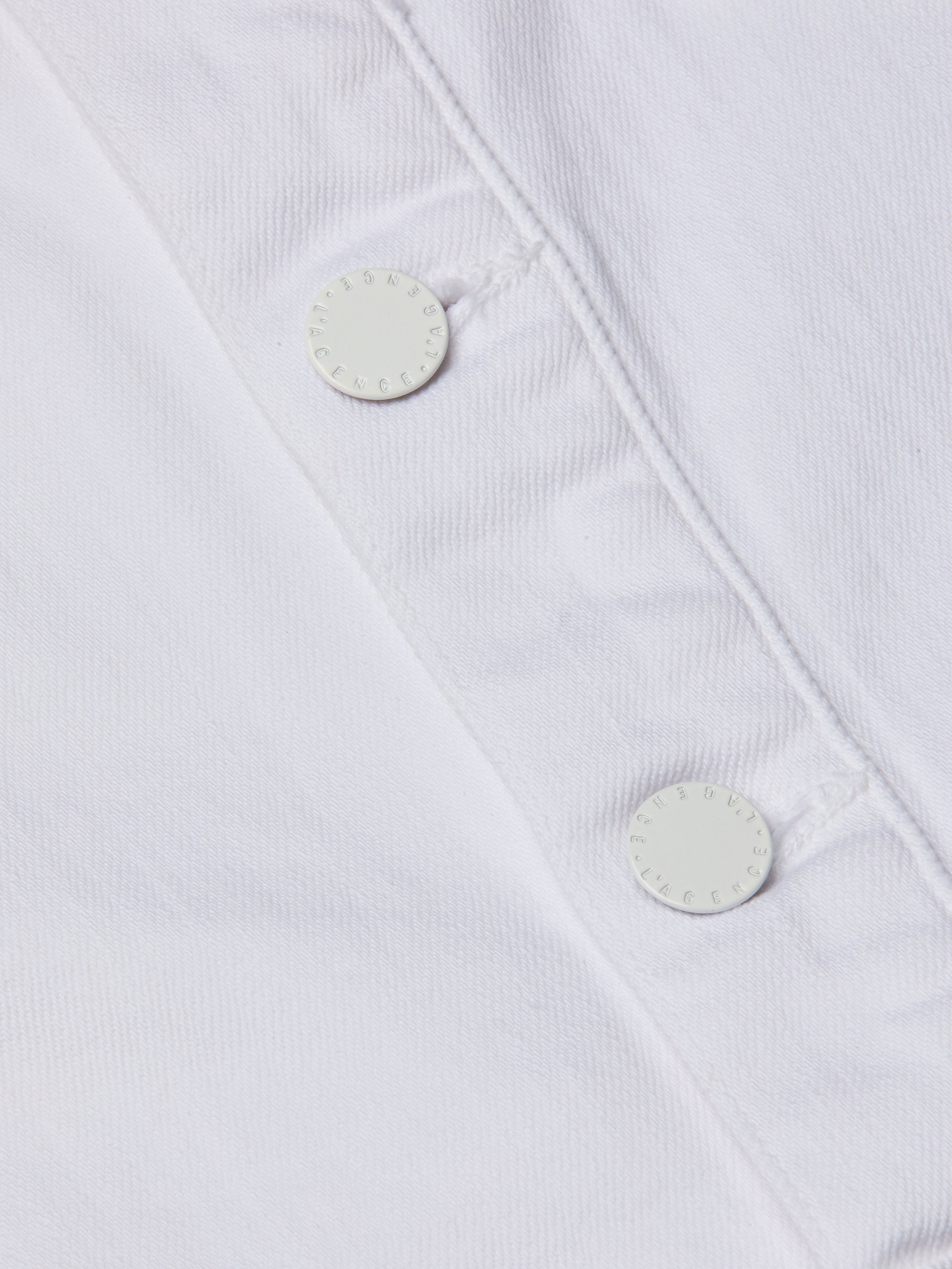 Close-up of the Kirstie Jean Shirt Jacket’s white denim fabric, highlighting two white round buttons with neatly stitched buttonholes and a subtly textured appearance.
