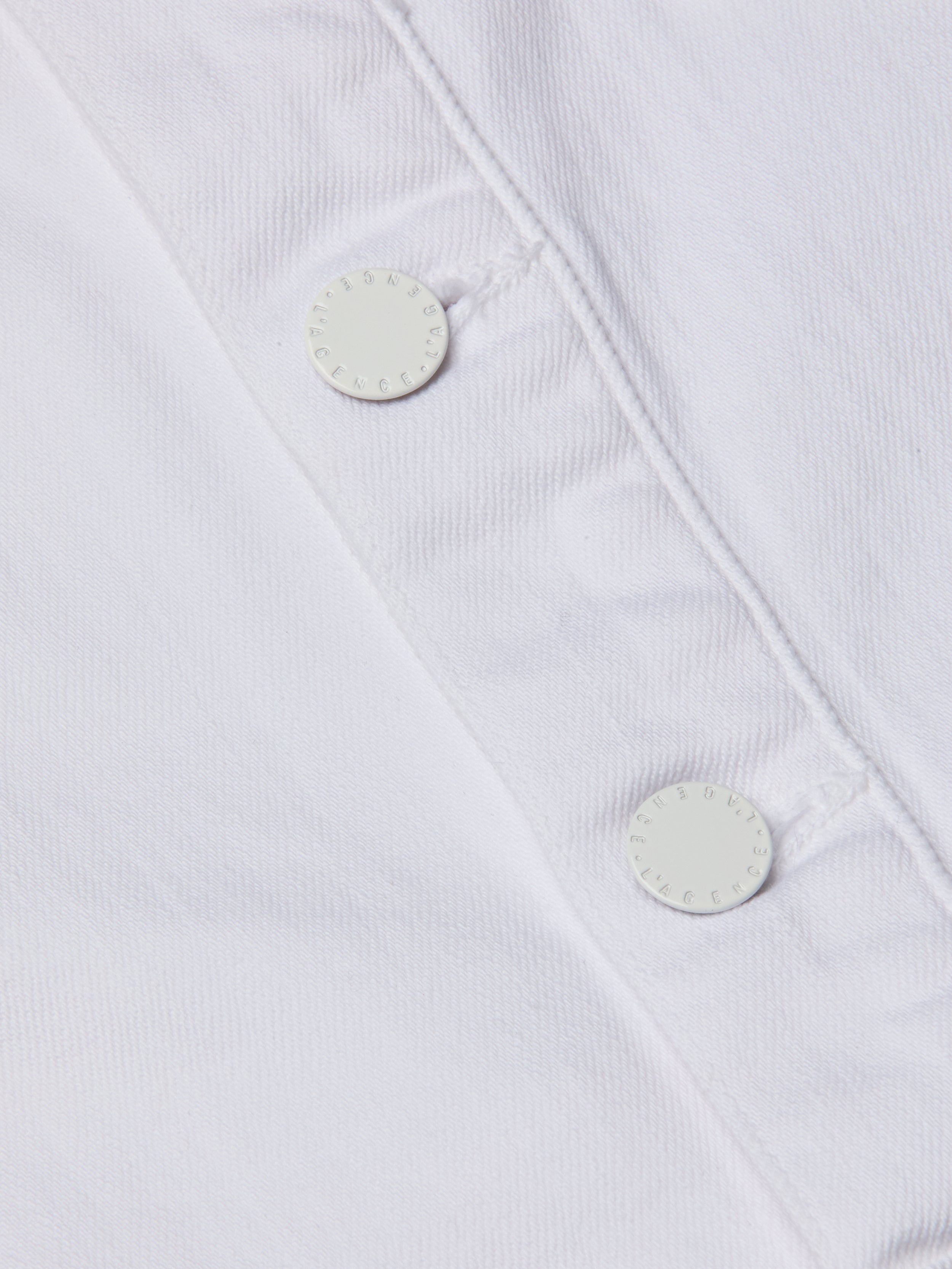 Close-up of the Kirstie Jean Shirt Jacket’s white denim fabric, highlighting two white round buttons with neatly stitched buttonholes and a subtly textured appearance.