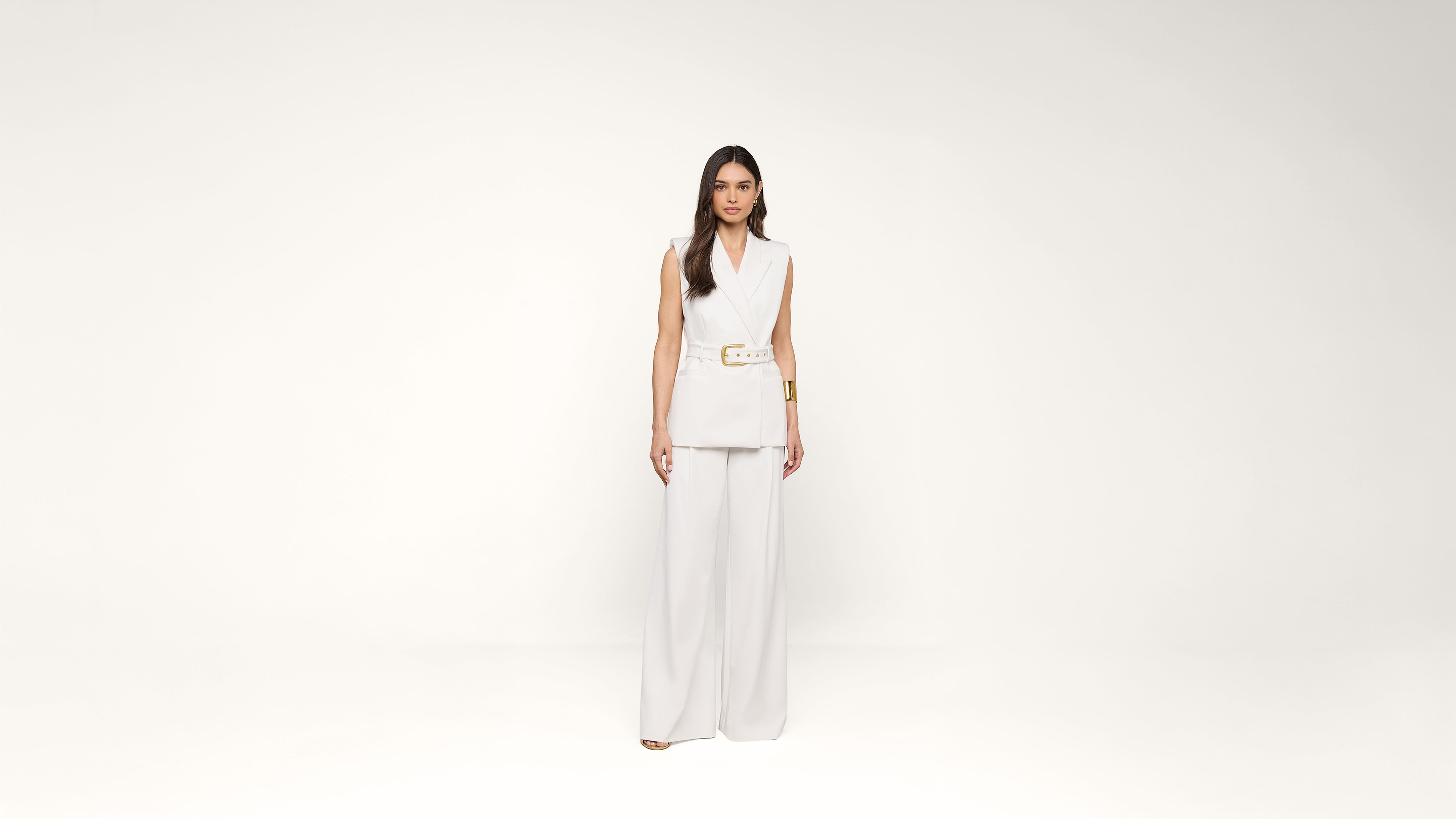 A woman stands against a plain white background, wearing a sleeveless white blazer with a belt and wide-legged white pants. She has long dark hair and a neutral expression.