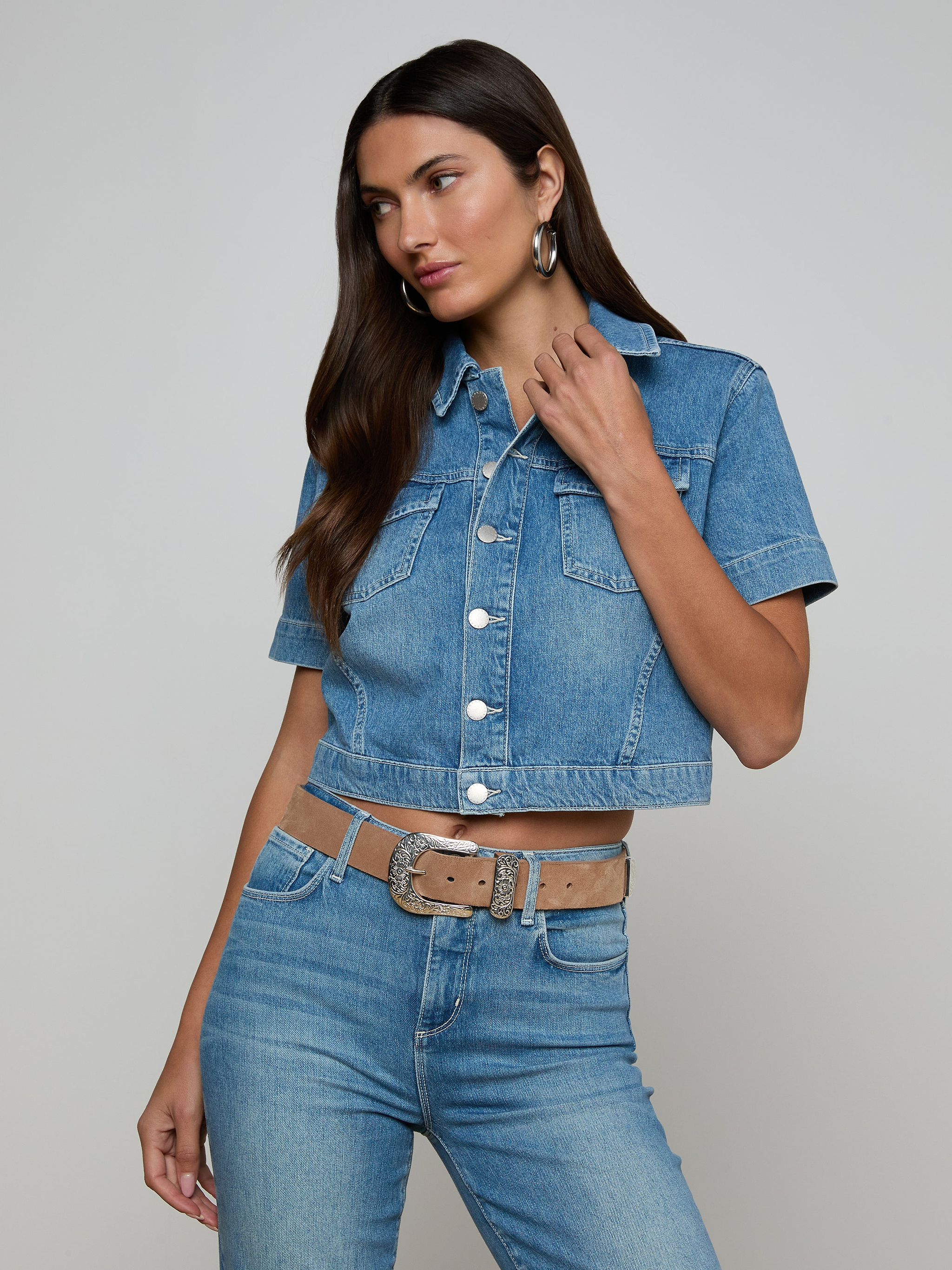 Wearing the Kirstie Jean Shirt Jacket, a woman with long dark hair pairs the short-sleeve cropped denim top with matching high-waisted jeans, a tan belt, and hoop earrings, standing against a plain light background.