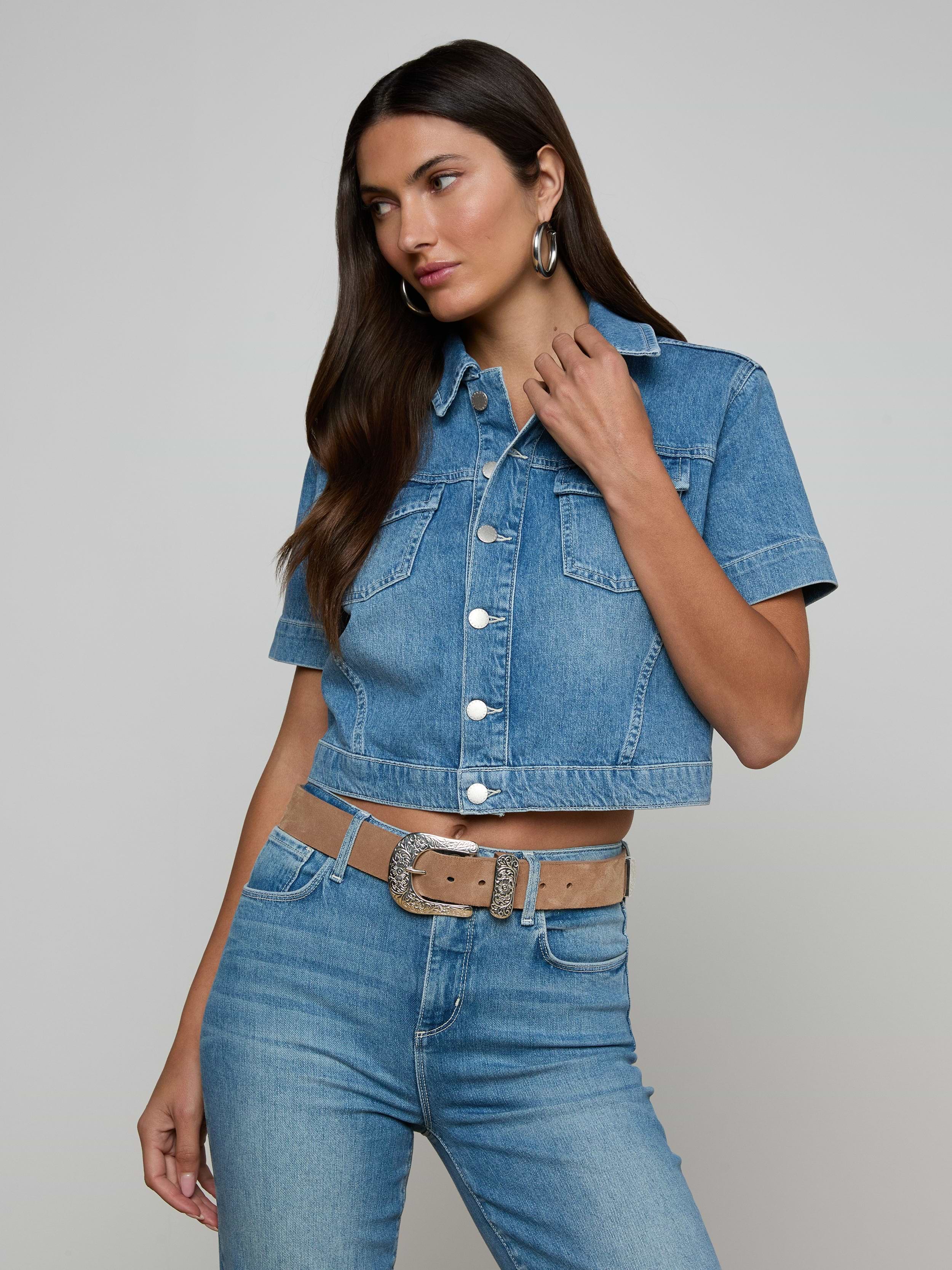 Wearing the Kirstie Jean Shirt Jacket, a woman with long dark hair pairs the short-sleeve cropped denim top with matching high-waisted jeans, a tan belt, and hoop earrings, standing against a plain light background.