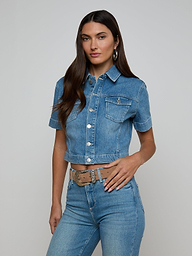 Wearing the Kirstie Jean Shirt Jacket—a cropped denim piece with short sleeves and front pockets—paired with high-waisted blue jeans, a woman stands against a plain light background.