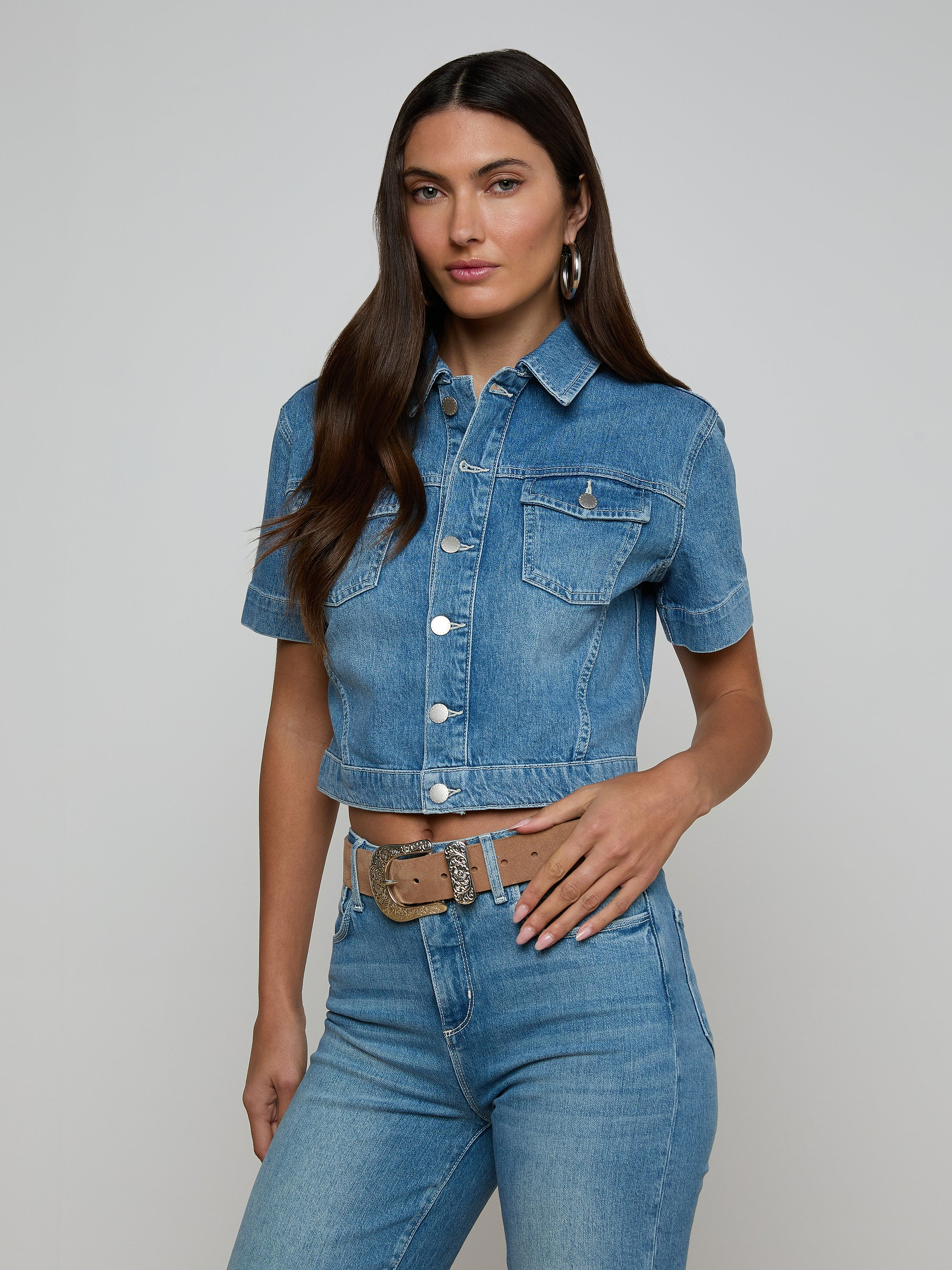 Wearing the Kirstie Jean Shirt Jacket—a cropped denim piece with short sleeves and front pockets—paired with high-waisted blue jeans, a woman stands against a plain light background.