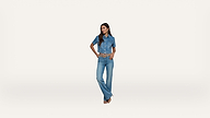 A woman stands against a white background wearing a short-sleeve denim button-up shirt and flared jeans. She has long dark hair and one hand on her hip.
