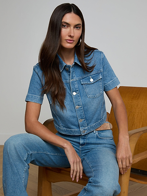 A woman with long brown hair confidently looks at the camera while sitting on a wooden chair, wearing the Kirstie Jean Shirt Jacket with matching jeans against a plain background.