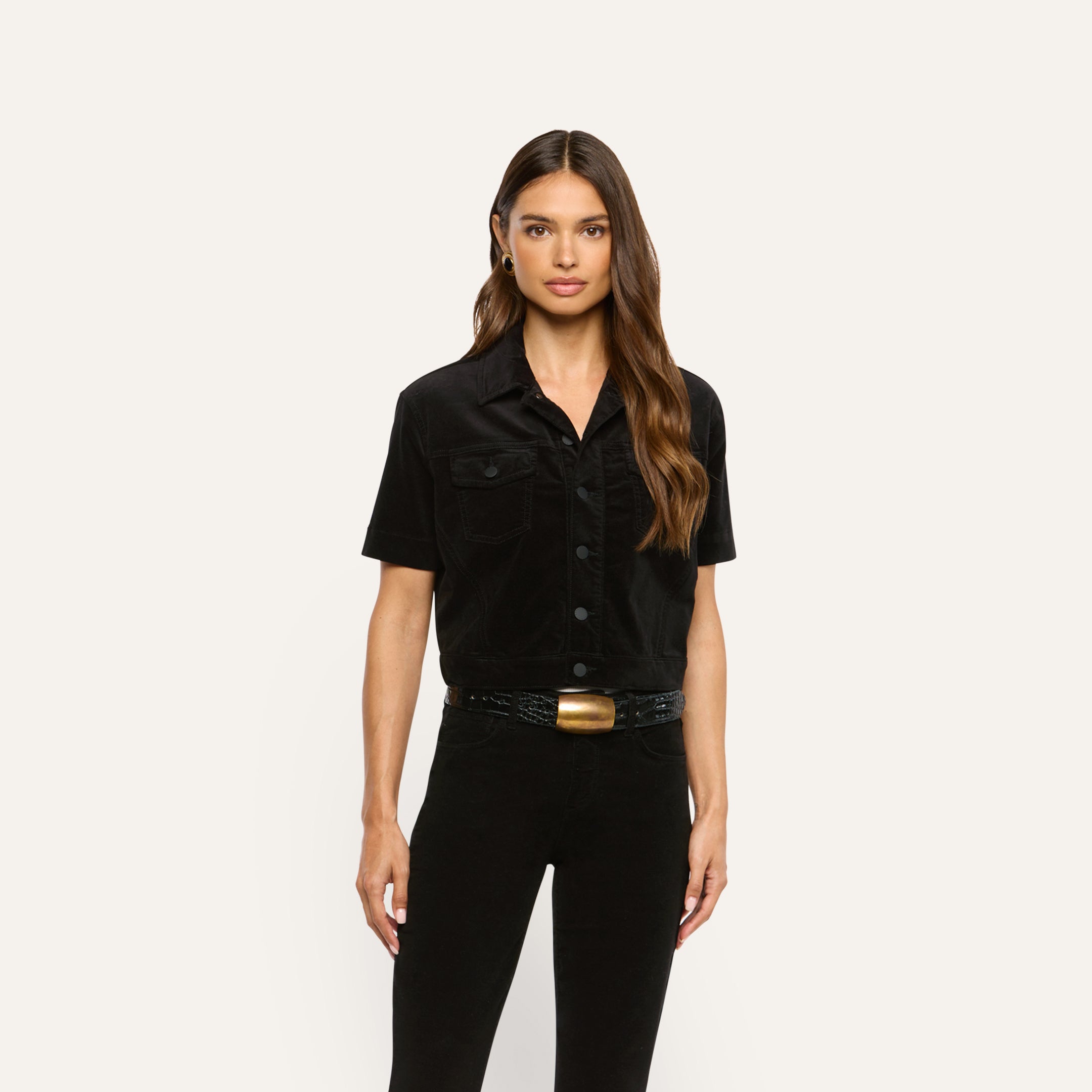A woman wears a black Kirstie Velvet Shirt Jacket with chest pockets and a black belt with a gold buckle over black pants.