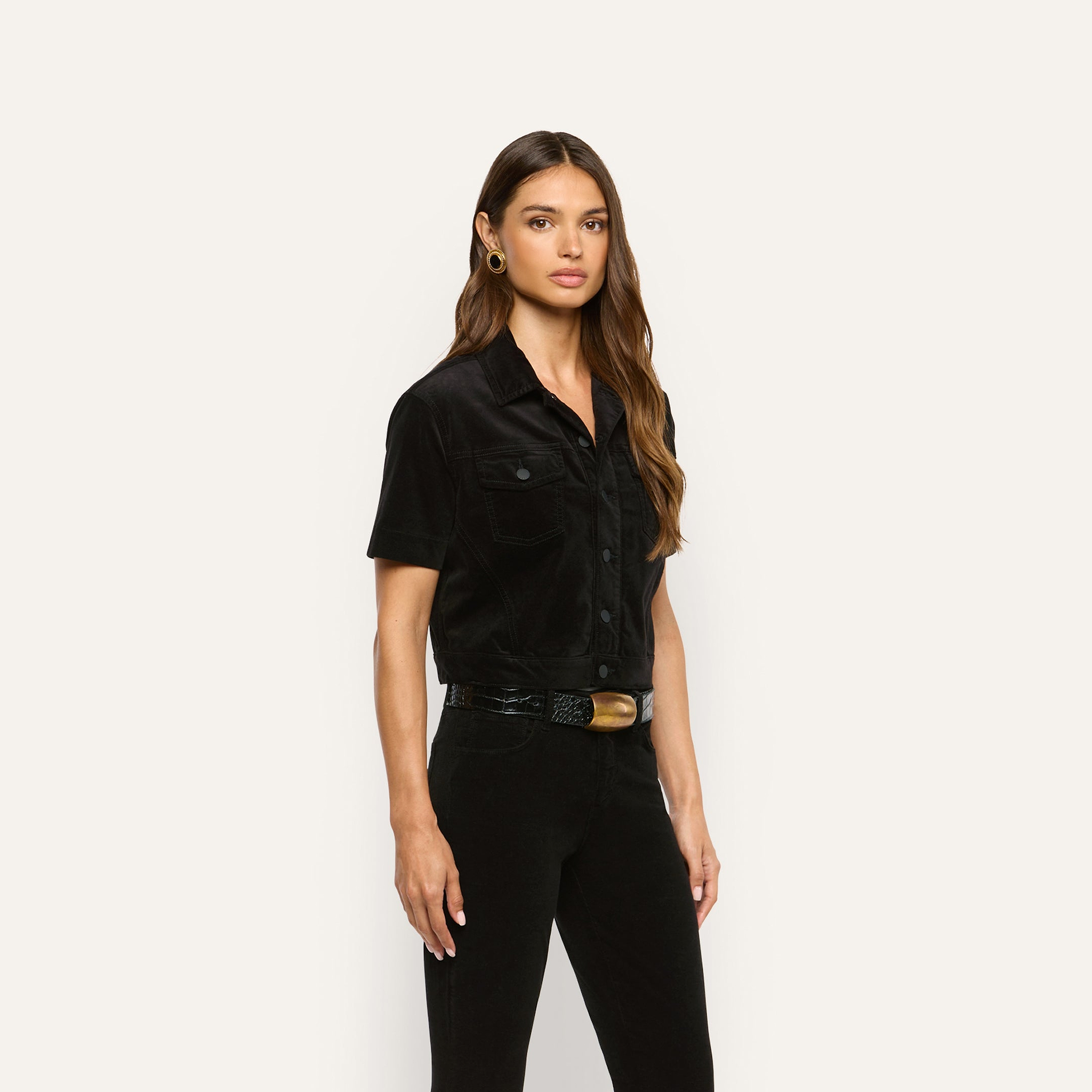 A woman wears a black short-sleeve Kirstie Velvet Shirt Jacket with front flap pockets and black pants with a wide-buckle belt.