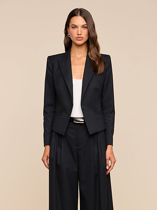 A woman with long, wavy brown hair and a neutral expression stands facing forward against a beige background, wearing the Misa Blazer—a single-breasted pinstripe blazer—paired with wide-leg pants over a white top.