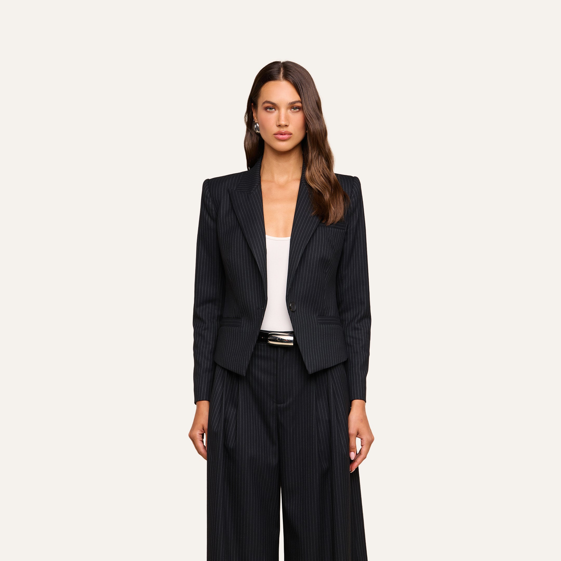 A woman with long, wavy brown hair and a neutral expression stands facing forward against a beige background, wearing the Misa Blazer—a single-breasted pinstripe blazer—paired with wide-leg pants over a white top.