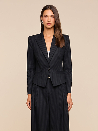 A woman with long brown hair wears the Misa Blazer—a dark pinstripe, single-breasted piece—paired with matching wide-leg trousers, standing against a beige background and facing forward with a neutral expression. The blazer lends vintage-inspired satin elegance to her sophisticated look.