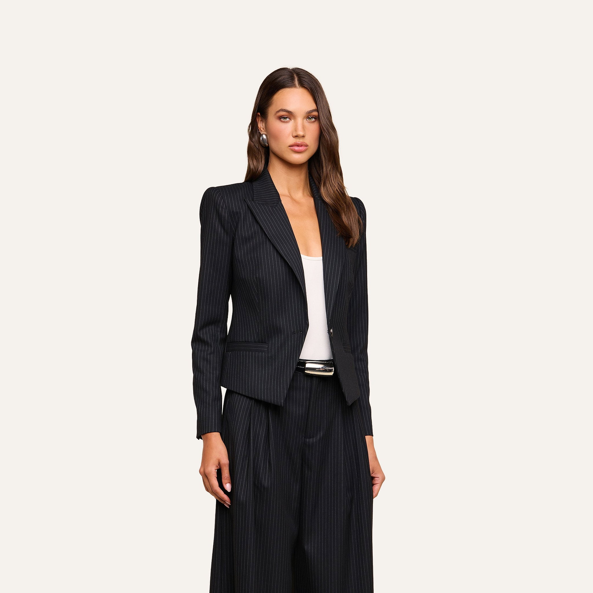 A woman stands against a plain beige background, looking forward with a neutral expression. She wears the Misa Blazer—a sleek, single-breasted black pinstripe jacket with a vintage-inspired satin finish—paired with matching wide-leg pants over a white top, creating an effortlessly polished look.
