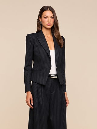 A woman stands against a plain beige background, looking forward with a neutral expression. She wears the Misa Blazer—a sleek, single-breasted black pinstripe jacket with a vintage-inspired satin finish—paired with matching wide-leg pants over a white top, creating an effortlessly polished look.