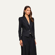 A woman stands against a beige background wearing the Misa Blazer, a black single-breasted pinstripe jacket, paired with matching wide-leg pants. She has long brown hair, silver hoop earrings, and a calm expression.