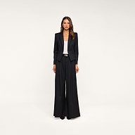 A woman stands facing forward against a plain beige background, wearing the Misa Blazer—a black single-breasted pinstripe blazer—paired with wide-leg trousers, a white top, and black pointed shoes.
