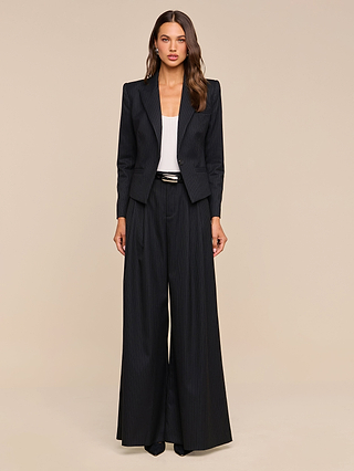 A woman stands facing forward against a plain beige background, wearing the Misa Blazer—a black single-breasted pinstripe blazer—paired with wide-leg trousers, a white top, and black pointed shoes.