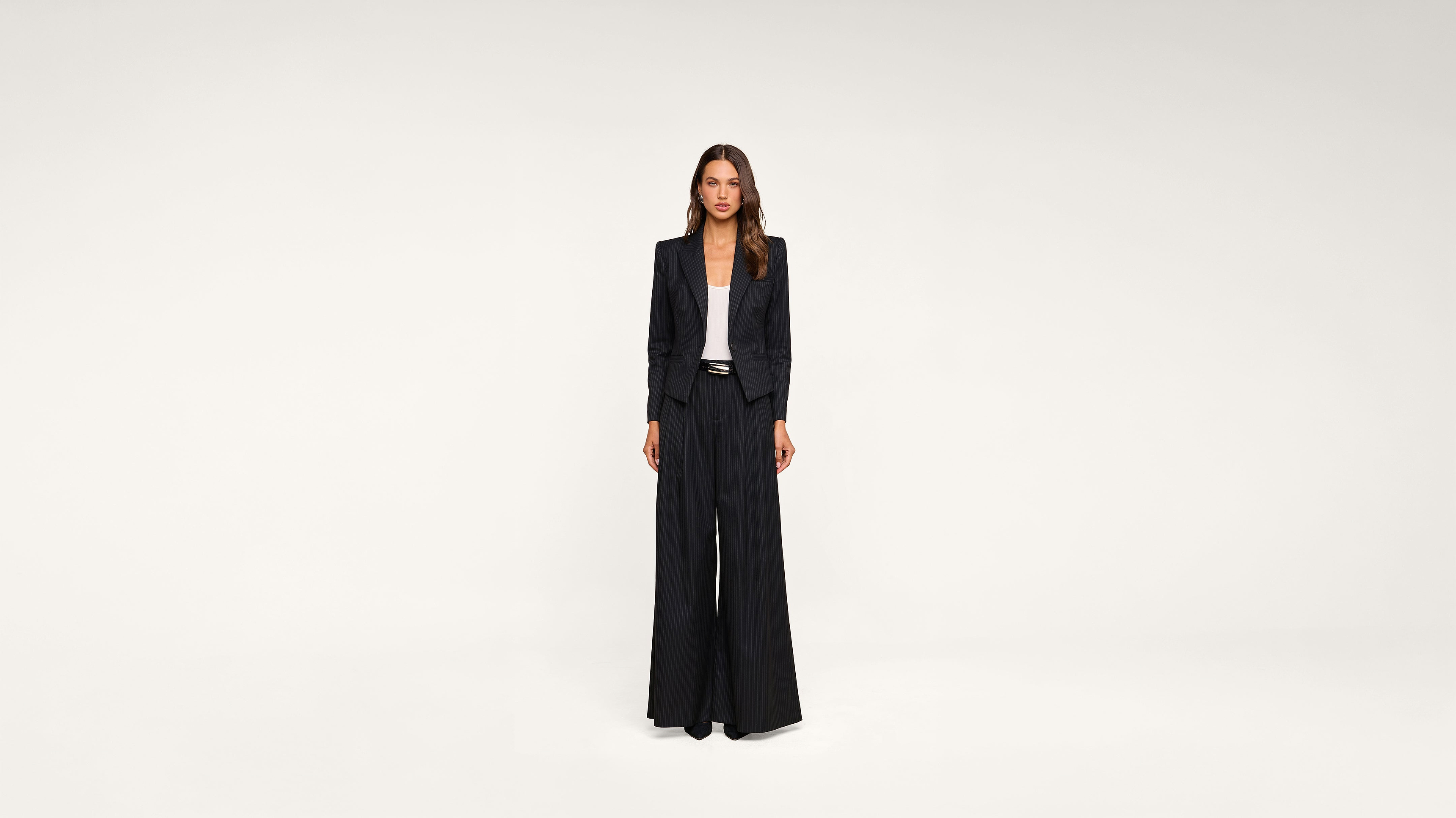 A woman stands against a plain white background, wearing a black blazer, a white top, and wide-leg black trousers, with her hands by her sides.