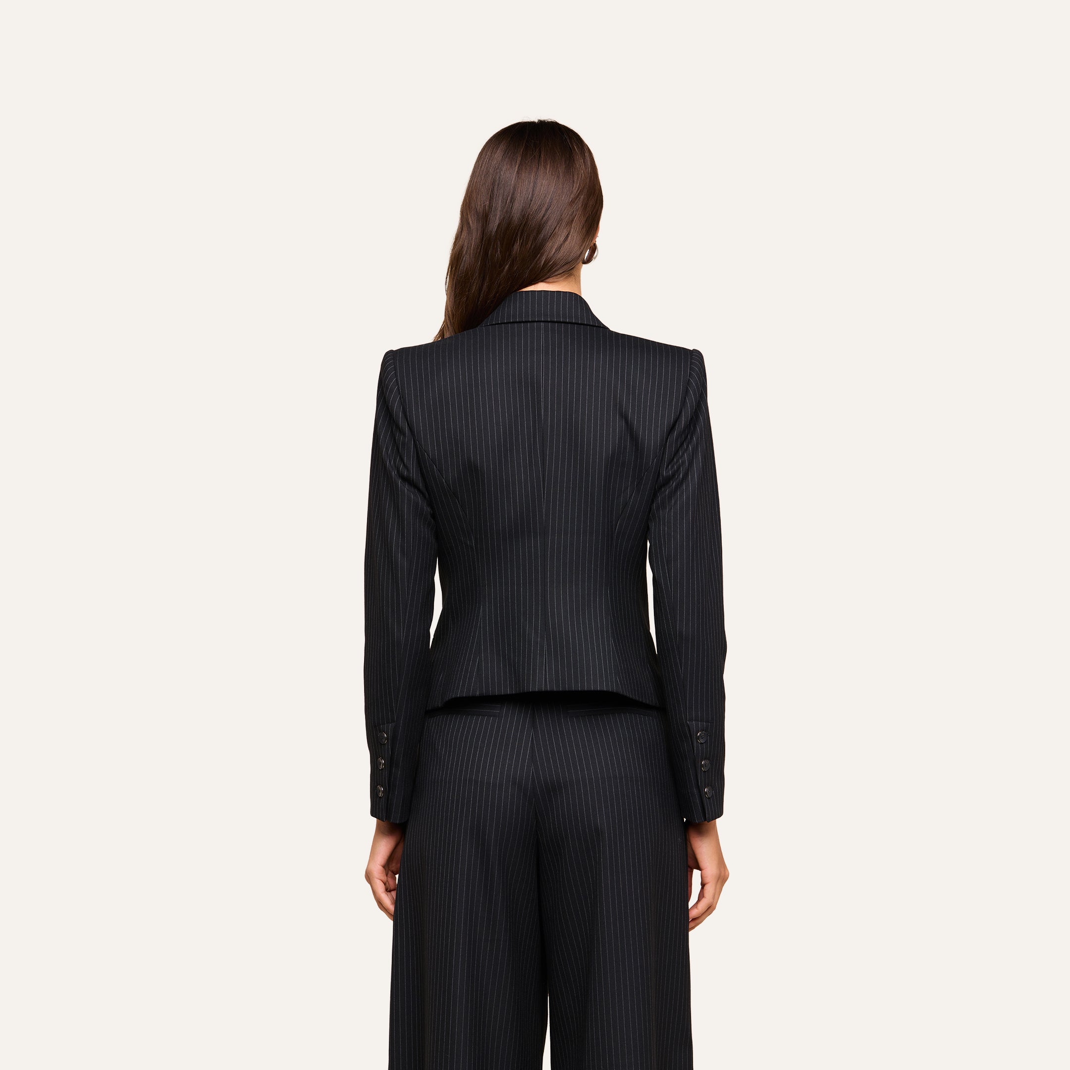 A woman with long brown hair stands facing away from the camera against a plain beige background, modeling the black pinstripe Misa Blazer.