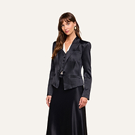 A woman stands against a plain background, wearing the black pinstripe Misa Blazer with a matching vest and a long black skirt. Her ensemble features elements of satin suiting and is complemented by a crisp white top. She has long brown hair with bangs and is looking at the camera.