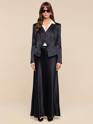 A woman in sunglasses wears the Misa Blazer—a black pinstripe single-breasted blazer—paired with a matching vest, long satin skirt, and white collared shirt, standing against a beige background.