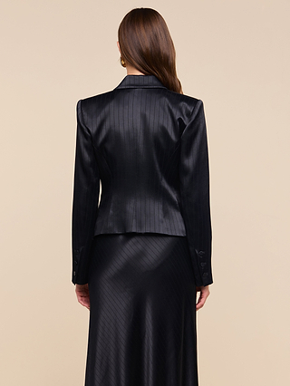 A woman with long brown hair is shown from behind, wearing the black pinstripe satin Misa Blazer and a matching skirt against a plain beige background.