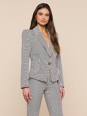A woman with long, straight brown hair and a neutral expression stands against a beige background, wearing the Misa Seersucker Blazer featuring black and white vertical stripes and matching pants.