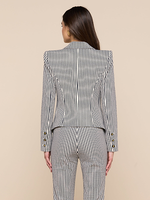 A woman with long brown hair is seen from behind wearing the Misa Seersucker Blazer, featuring stylish black and white stripes, paired with matching pants against a plain beige background.