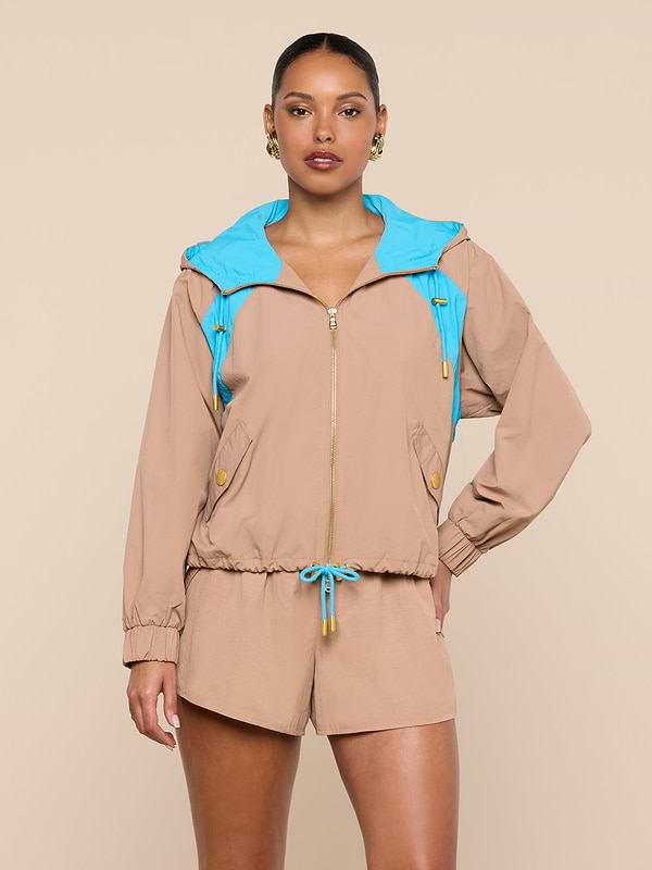 A woman stands with one hand on her hip, wearing the Lark Windbreaker by L’AGENCE, a tan jacket with retro color blocking, a bright blue hood, front zipper, drawstring waist, and yellow buttons. She pairs it with matching tan shorts and gold earrings, her hair tied back.