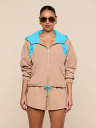 A person stands facing forward against a plain white background, wearing sunglasses and the Lark Windbreaker—a beige jacket with blue accents featuring retro color blocking—paired with matching beige shorts.