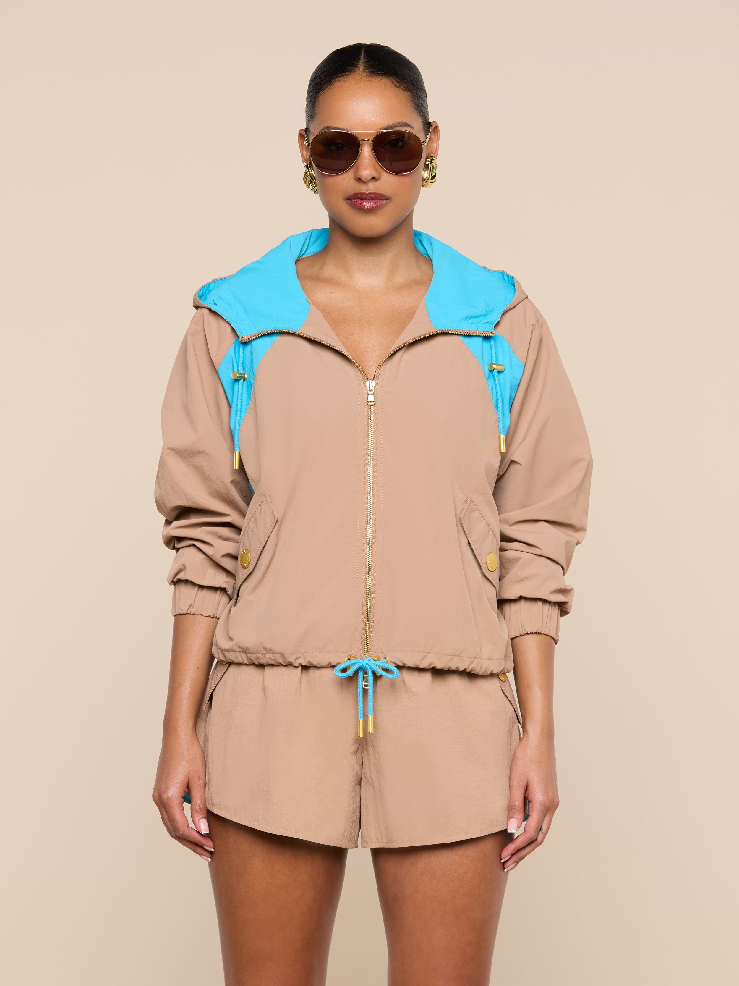 A person stands facing forward against a plain white background, wearing sunglasses and the Lark Windbreaker—a beige jacket with blue accents featuring retro color blocking—paired with matching beige shorts.