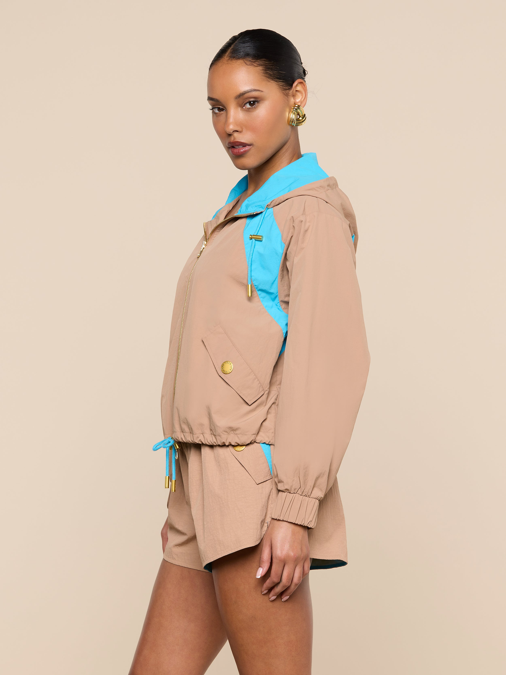 A woman stands sideways wearing the L’AGENCE Lark Windbreaker in beige with blue retro color blocking, paired with matching beige shorts. She accessorizes with gold hoop earrings and has her hair pulled back, posing against a plain light background.