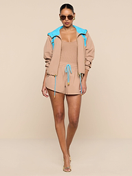 A woman stands confidently against a plain white background, wearing the tan romper paired with the Lark Windbreaker—a retro color-blocking hooded jacket featuring blue lining—along with sunglasses and heels.