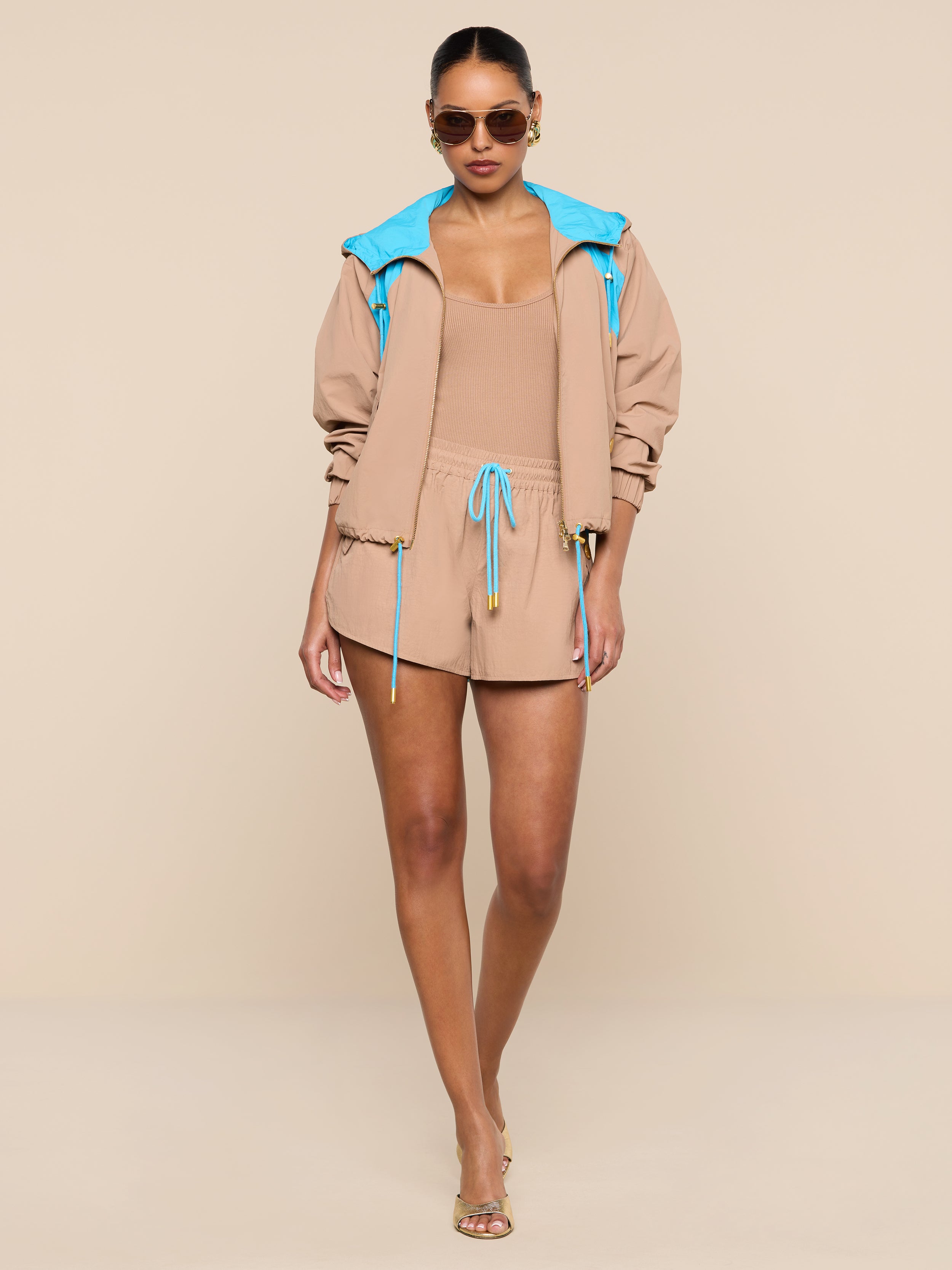 A woman stands confidently against a plain white background, wearing the tan romper paired with the Lark Windbreaker—a retro color-blocking hooded jacket featuring blue lining—along with sunglasses and heels.