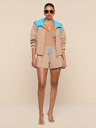 A woman stands confidently facing forward against a plain white background, wearing the Grayson Nylon Short paired with a tan crepe nylon jacket featuring a blue hood and a matching tan top. She accessorizes her look with sunglasses and clear heels.