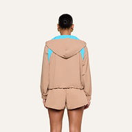 A person, shown from behind, is wearing the Lark Windbreaker—a tan hooded jacket with light blue accents and retro color blocking—paired with matching tan shorts. Their hair is styled in a bun and they accessorize with gold hoop earrings. The background is plain and light-colored.