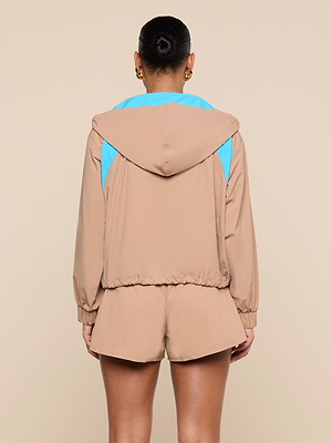 Une personne se tient debout, de face, vêtue du coupe-vent Lark, en tissu beige avec des couleurs bleues rétro sur les épaules, associé à un short beige assorti. Ses cheveux sont coiffés en chignon et elle porte des boucles doreilles dorées sur un fond beige uni.