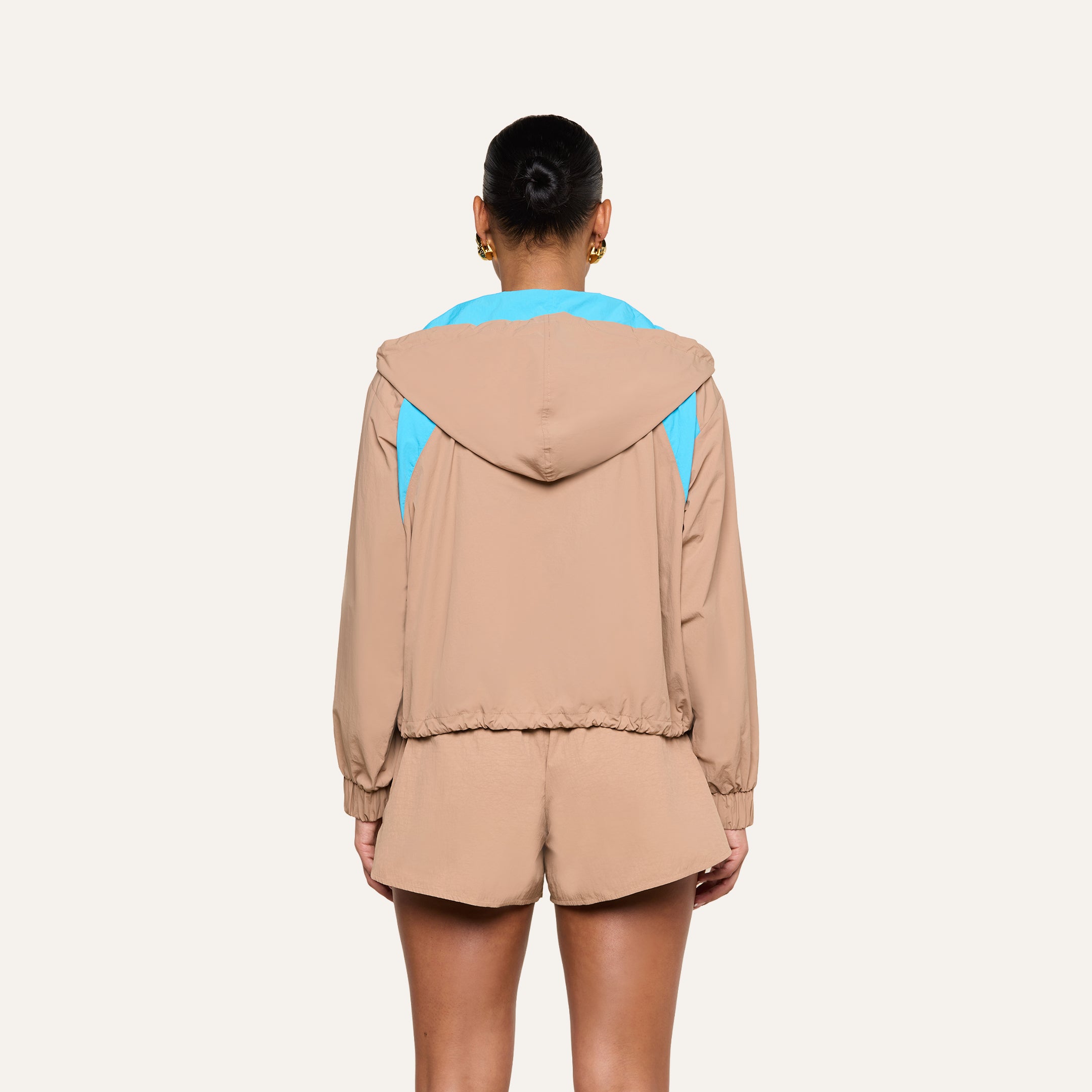 A person, shown from behind, is wearing the Lark Windbreaker—a tan hooded jacket with light blue accents and retro color blocking—paired with matching tan shorts. Their hair is styled in a bun and they accessorize with gold hoop earrings. The background is plain and light-colored.