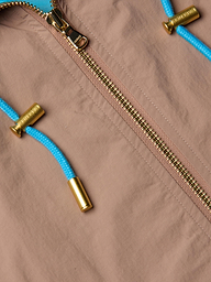 Close-up of the Lark Windbreaker by L’AGENCE in light brown, featuring a gold zipper and bright blue drawstrings tipped with metallic gold caps. The water-repellent fabric showcases subtle retro color blocking for a stylish touch.