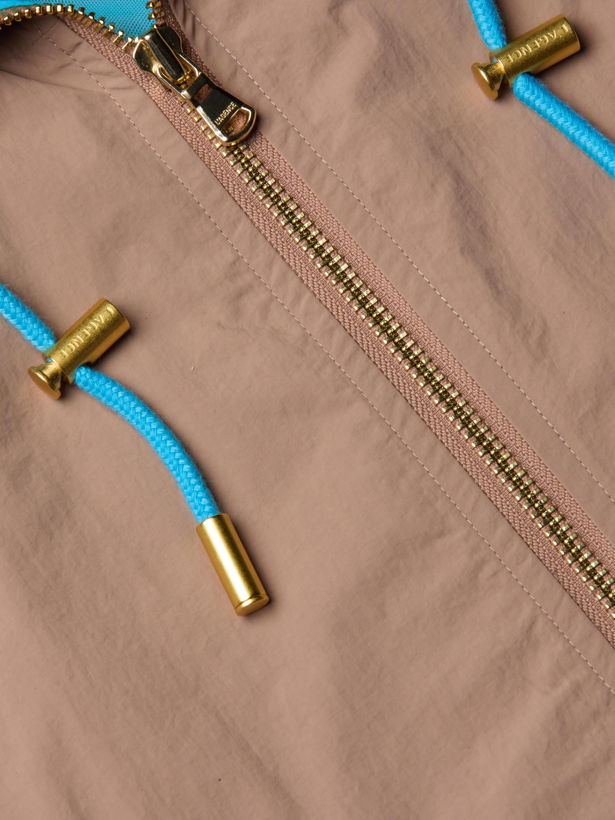 Close-up of the Lark Windbreaker by L’AGENCE in light brown, featuring a gold zipper and bright blue drawstrings tipped with metallic gold caps. The water-repellent fabric showcases subtle retro color blocking for a stylish touch.