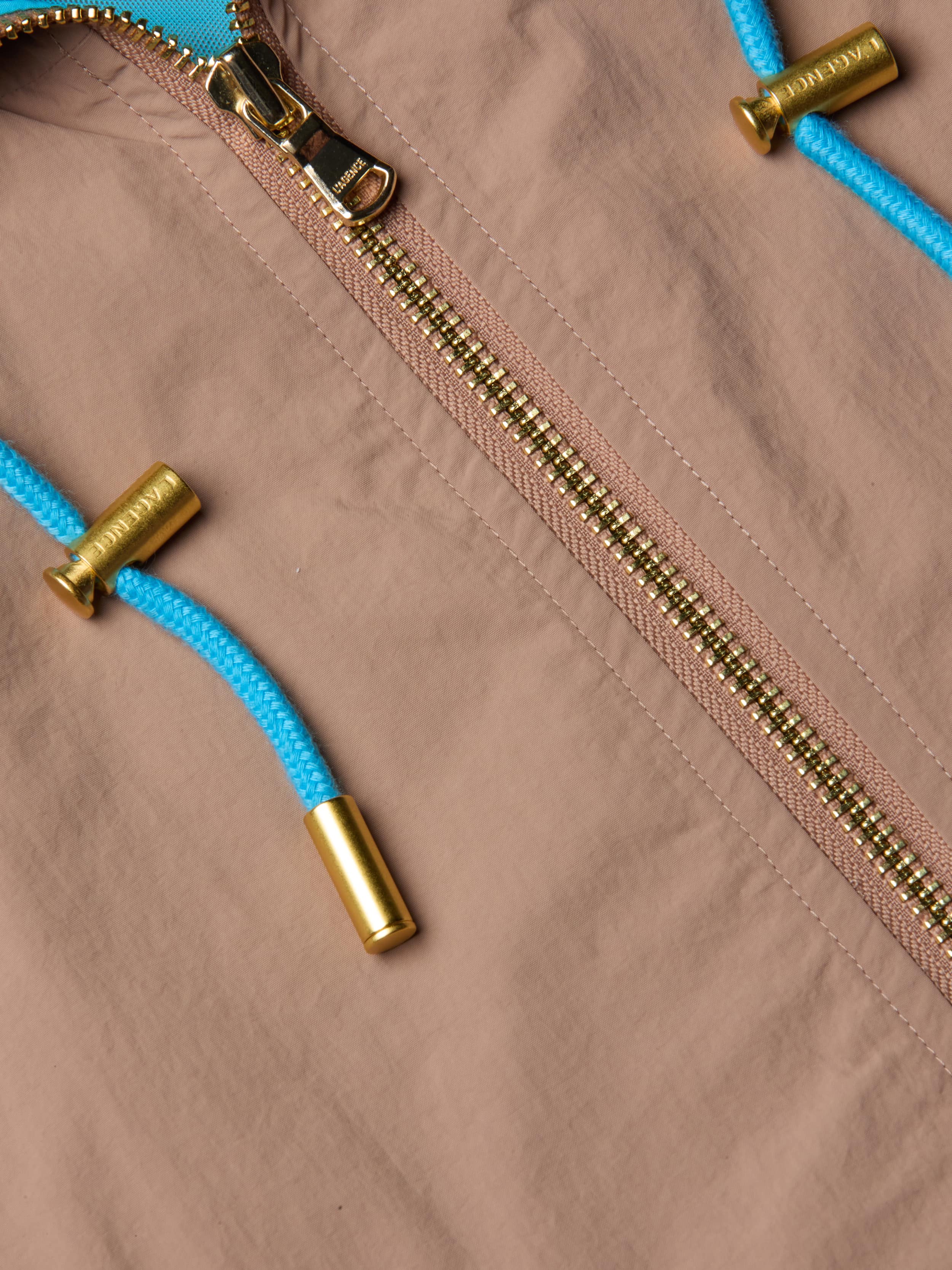 Close-up of the Lark Windbreaker by L’AGENCE in light brown, featuring a gold zipper and bright blue drawstrings tipped with metallic gold caps. The water-repellent fabric showcases subtle retro color blocking for a stylish touch.
