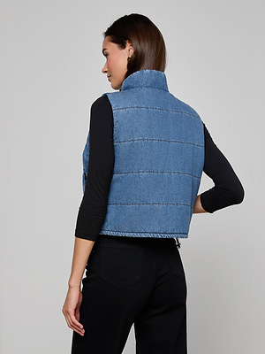 A woman with straight brown hair stands with her back to the camera against a light gray background, wearing the indigo Rola Puffer Vest in a cropped length over a black long-sleeve top and black pants.