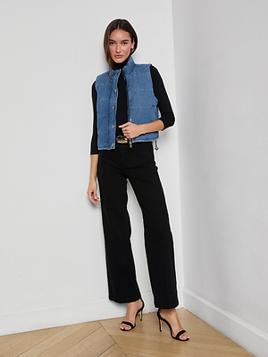 A woman with straight brown hair and a neutral expression stands indoors by a white wall, wearing the indigo Rola Puffer Vest over a black long-sleeve top, paired with black trousers and high-heeled sandals.