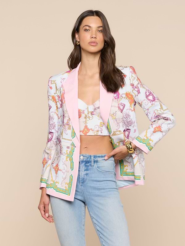 A woman with long brown hair poses against a beige background, wearing the Gweny Blazer—a colorful satin back crepe baroque toile blazer with pink trim—paired with a matching crop top, light blue jeans, gold earrings, and a bracelet.
