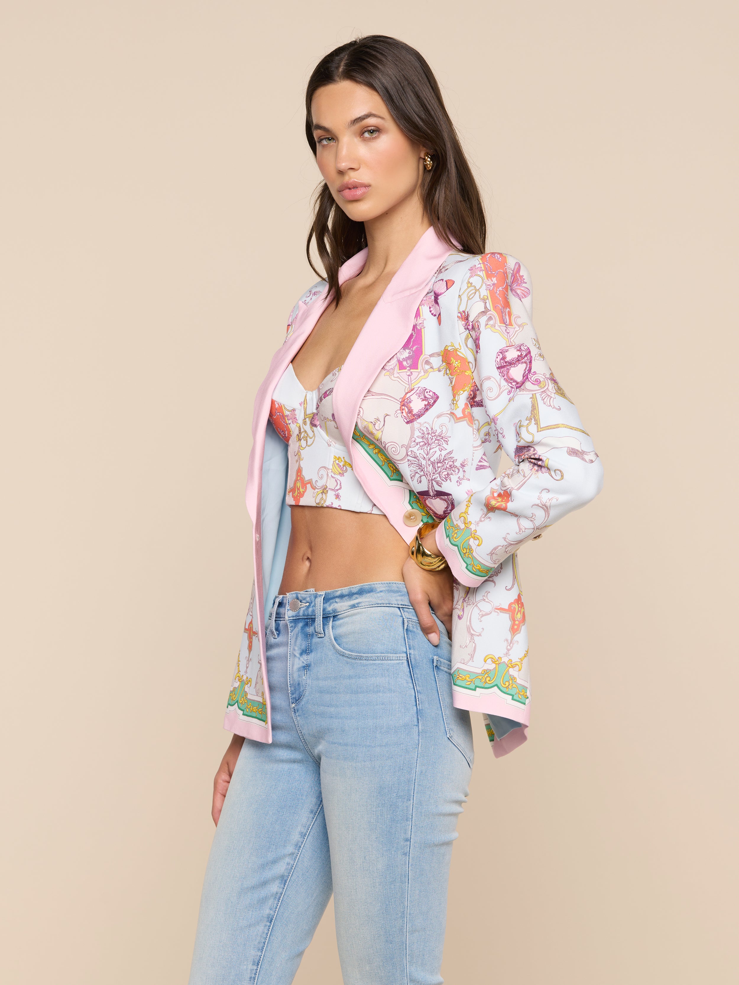 A woman poses in light blue jeans and a matching printed bustier top, styled with the Gweny Blazer featuring pastel pink accents, against a neutral beige background.