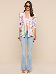 A woman stands against a beige background wearing sunglasses, the Gweny Blazer with pink lapels over a matching crop top, light blue flared jeans, and beige open-toe heels.
