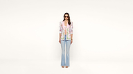 A woman wearing sunglasses, a colorful floral blazer, pink shirt, light blue flared jeans, and sandals stands against a plain white background, looking forward with her hands by her sides.