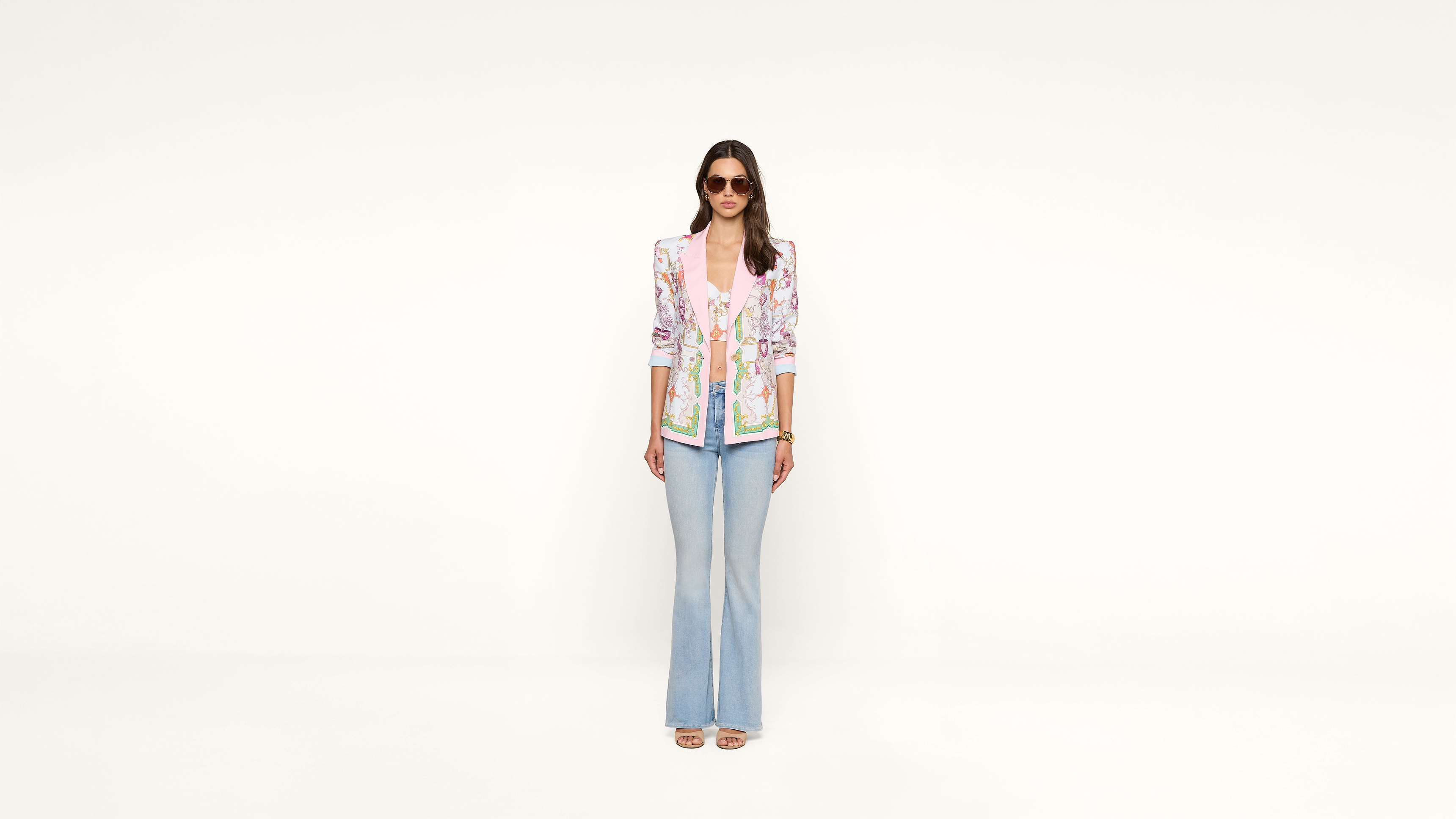 A woman wearing sunglasses, a colorful floral blazer, pink shirt, light blue flared jeans, and sandals stands against a plain white background, looking forward with her hands by her sides.