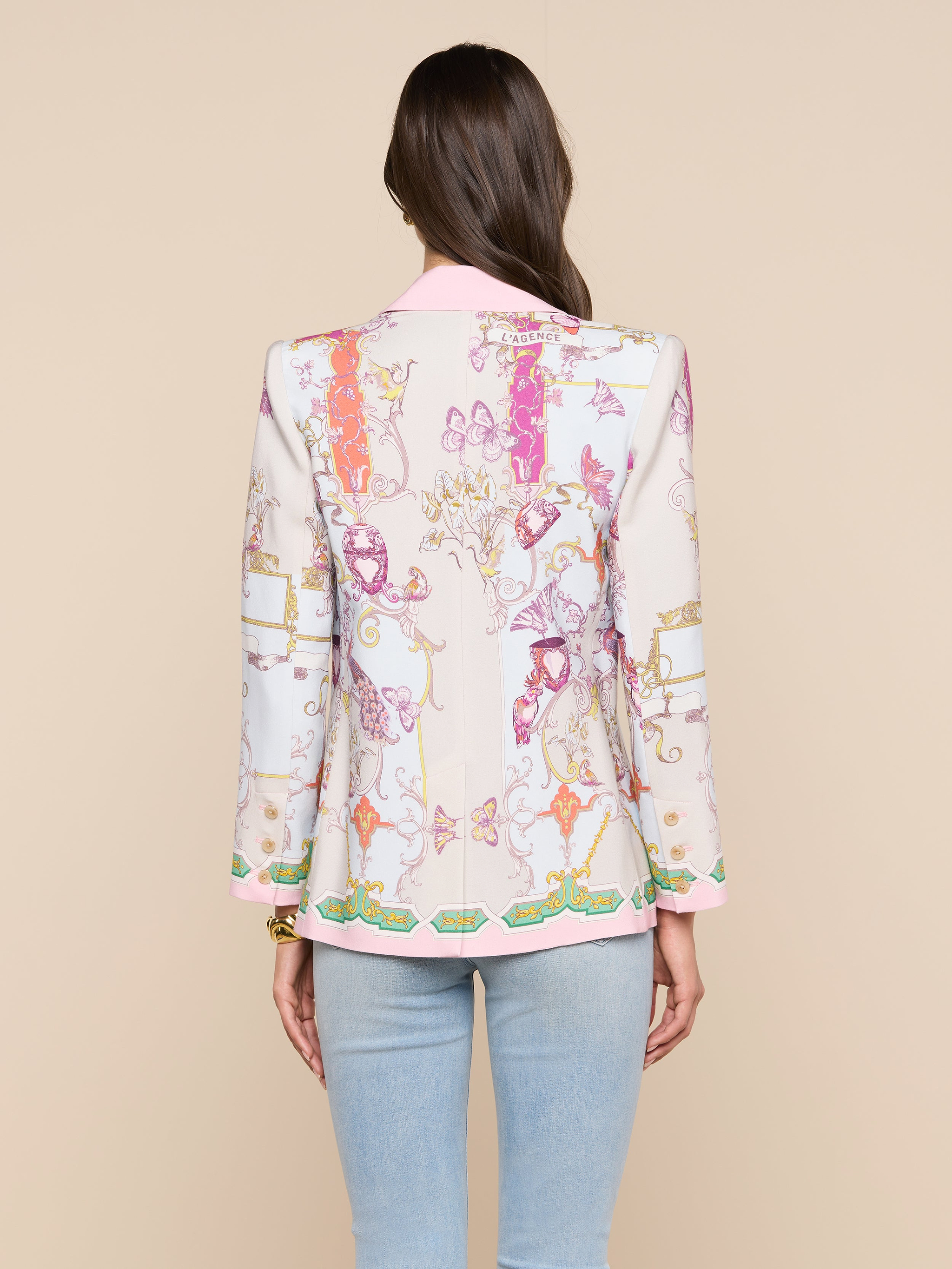 A woman with long brown hair stands with her back to the camera against a beige background, wearing the Gweny Blazer—a relaxed fit blazer with colorful patterns and pink trim—paired with light blue jeans.
