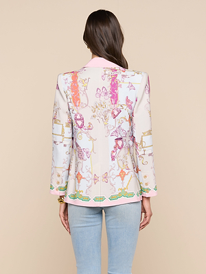 A woman with long brown hair stands with her back to the camera against a beige background, wearing the Gweny Blazer—a relaxed fit blazer with colorful patterns and pink trim—paired with light blue jeans.